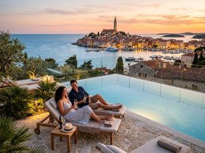 High-definition realistic travel photograph of an Israeli couple relaxing beside a luxurious infinity pool at sunset, overlooking Rovinj’s glowing stone old town, marina, and Adriatic Sea; elegant modern hotel terraces, natural warm light, cinematic depth of field, no text.