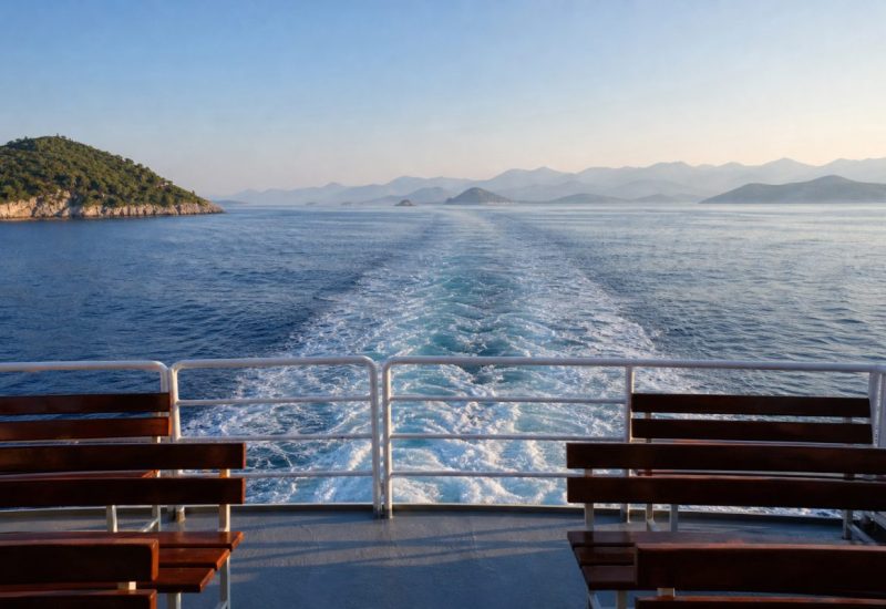 View from the open stern deck of a passenger boat crossing the Adriatic, the white wake trailing across deep blue water toward layered Dalmatian islands fading into soft haze on the horizon, a simple metal railing and empty wooden benches in the foreground, calm sea with gentle swell, clear early morning light and a pale sky. Use balanced, natural contrast with a realistic tonal range. Preserve fine detail throughout the image, with smooth highlight roll-off, detailed shadows, balanced exposure, and natural, accurate colors.