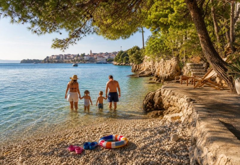 A warm late-afternoon scene in a small sheltered cove on the northern Adriatic coast of Croatia: pale limestone pebbles at the waterline, clear turquoise shallows fading into deeper blue, and low Mediterranean pines casting soft dappled shade over a stone terrace above the beach. In the middle distance a family with two young children wades in ankle-deep water, seen from behind, colourful water shoes and a striped inflatable ring resting on the pebbles beside them. Terracotta rooftops of a small coastal town sit low on the far headland. Use balanced, natural contrast with a realistic tonal range. Preserve fine detail throughout the image, with smooth highlight roll-off, detailed shadows, balanced exposure, and natural, accurate colors.