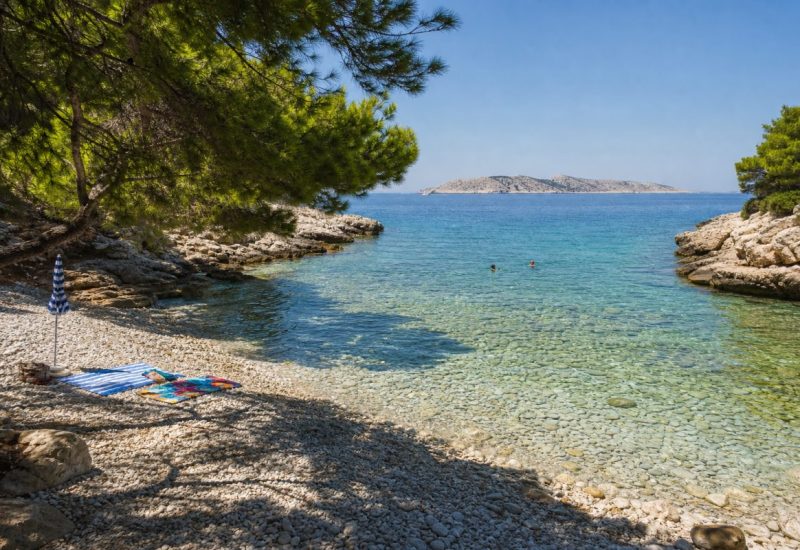 A wide, sunlit photograph of a Croatian Adriatic cove in late morning: a curving beach of smooth white limestone pebbles meeting turquoise, glass-clear water where the rocky seabed is visible through the shallows. Weathered pale karst rock ledges frame both sides of the bay, with old Aleppo pines leaning over the shore and casting dappled shade on the stones. A few colourful towels and a folded parasol rest on the pebbles, and two swimmers are in the water far from the camera, small in the frame. Distant limestone islands sit low on the horizon under a clear summer sky. Photorealistic travel photography, natural daylight, medium-wide lens. Use balanced, natural contrast with a realistic tonal range. Preserve fine detail throughout the image, with smooth highlight roll-off, detailed shadows, balanced exposure, and natural, accurate colors.