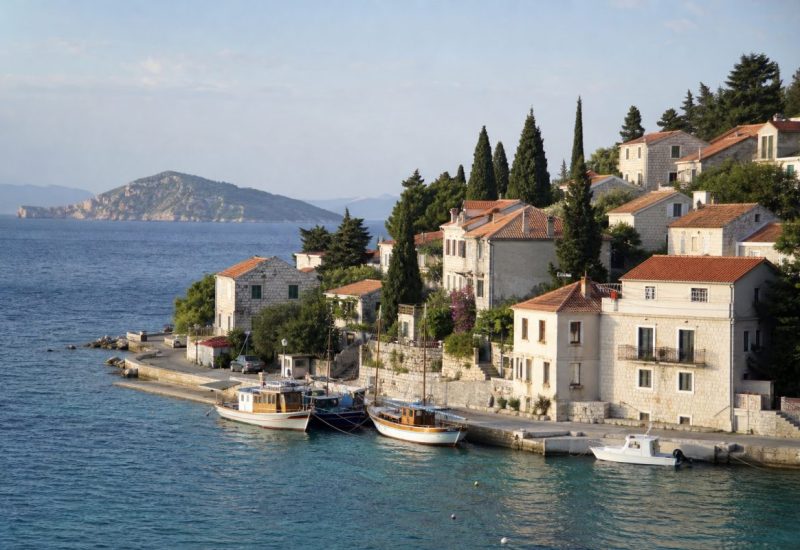 A wide daylight photograph of a small Dalmatian harbour town on the Adriatic coast in late summer: pale limestone houses with terracotta roofs stacked up a low hillside, a few wooden fishing boats and a small white motorboat moored along a stone quay, clear turquoise water in the foreground, tall dark green cypresses and pines between the houses, a rocky island silhouette on the horizon under a soft blue sky with light haze. Shot from a slightly elevated viewpoint across the water, calm and uncluttered, no people in the frame. Photographic realism, medium telephoto compression, soft late afternoon light. Use balanced, natural contrast with a realistic tonal range. Preserve fine detail throughout the image, with smooth highlight roll-off, detailed shadows, balanced exposure, and natural, accurate colors.