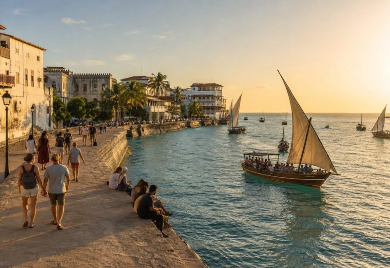 Wide elevated golden hour photograph of the Stone Town seafront in Zanzibar, warm low sunlight across the water, a group of relaxed international travelers in light summer clothing strolling along the stone waterfront walkway and sitting on the low sea wall, traditional wooden dhow sailing boats out on the turquoise water with people aboard, weathered coral stone buildings and palm trees framing the background, soft haze over the Indian Ocean, candid documentary travel photography feel, people small within a spacious composition. Use balanced, natural contrast with a realistic tonal range. Preserve fine detail throughout the image, with smooth highlight roll-off, detailed shadows, balanced exposure, and natural, accurate colors.
