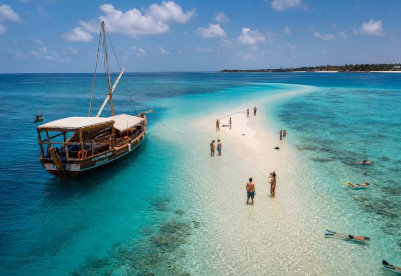 Wide aerial travel photograph of a traditional wooden sailing dhow anchored beside a bright turquoise sandbank off the coast of Zanzibar. A small group of holidaymakers in swimwear stand in shallow ankle deep water beside the sandbank, some snorkeling nearby, others walking on the white sand strip. Deep blue ocean surrounds the pale sandbar, coral reef patches visible under clear water, tropical sky with soft scattered clouds, warm late morning light. The people are small in the frame but clearly relaxed and enjoying themselves. Use balanced, natural contrast with a realistic tonal range. Preserve fine detail throughout the image, with smooth highlight roll-off, detailed shadows, balanced exposure, and natural, accurate colors.