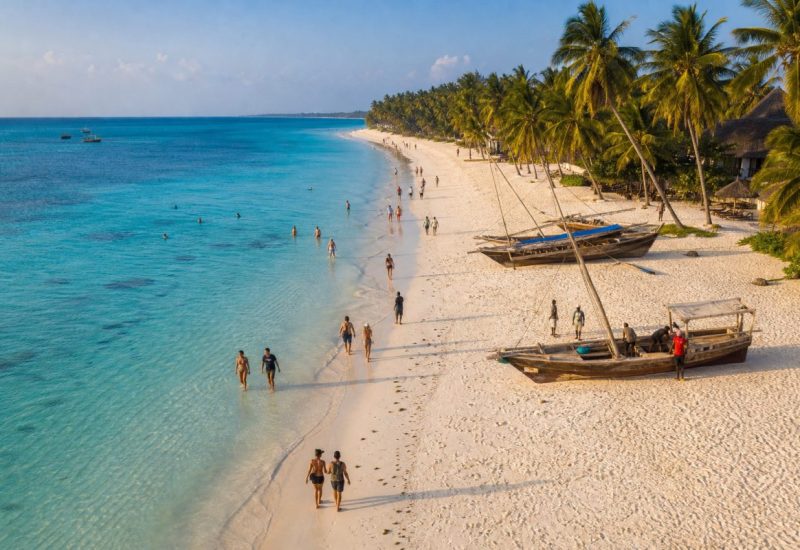 A wide, sunlit aerial view of a long white sand beach on the Indian Ocean coast of Zanzibar, turquoise shallows fading into deep blue water, small groups of holidaymakers walking along the waterline and wading in the shallow lagoon, a few swimmers further out, tall coconut palms leaning over the sand along the top of the frame, several traditional wooden sailing boats resting on the sand with people standing beside them, warm late afternoon light, calm and open composition, relaxed holiday atmosphere. Use balanced, natural contrast with a realistic tonal range. Preserve fine detail throughout the image, with smooth highlight roll-off, detailed shadows, balanced exposure, and natural, accurate colors.