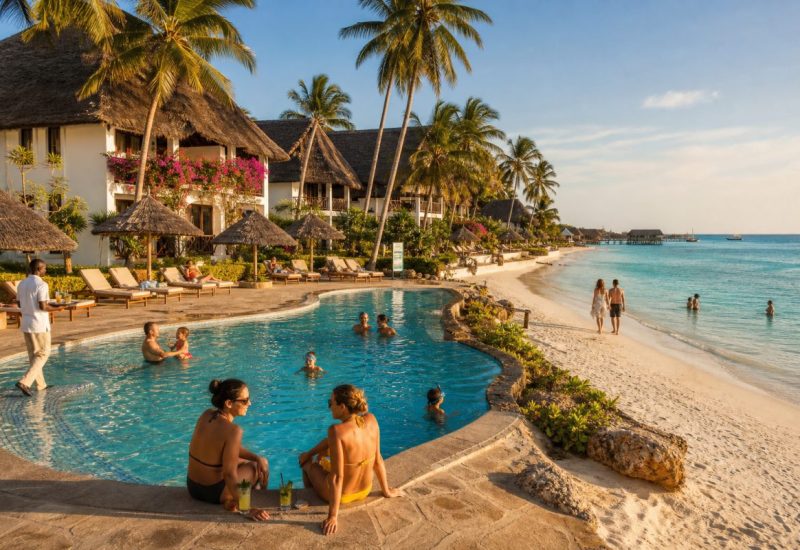 Wide sunlit view of a beachfront resort on Zanzibar in the late afternoon. In the foreground a freeform swimming pool edged with sun loungers and thatched parasols, where a family is swimming and two friends sit talking at the pool edge with fresh drinks. Behind the pool a row of low white resort buildings with palm thatch roofs sits among coconut palms and bougainvillea. Beyond them a wide strip of pale sand runs down to a bright turquoise lagoon, with a couple walking barefoot at the waterline and a few swimmers wading in the shallow clear water. A waiter in a light uniform carries a tray along the pool deck. Warm golden light, long soft shadows, relaxed holiday atmosphere, people appear naturally within the wide scene rather than posed. Use balanced, natural contrast with a realistic tonal range. Preserve fine detail throughout the image, with smooth highlight roll-off, detailed shadows, balanced exposure, and natural, accurate colors.